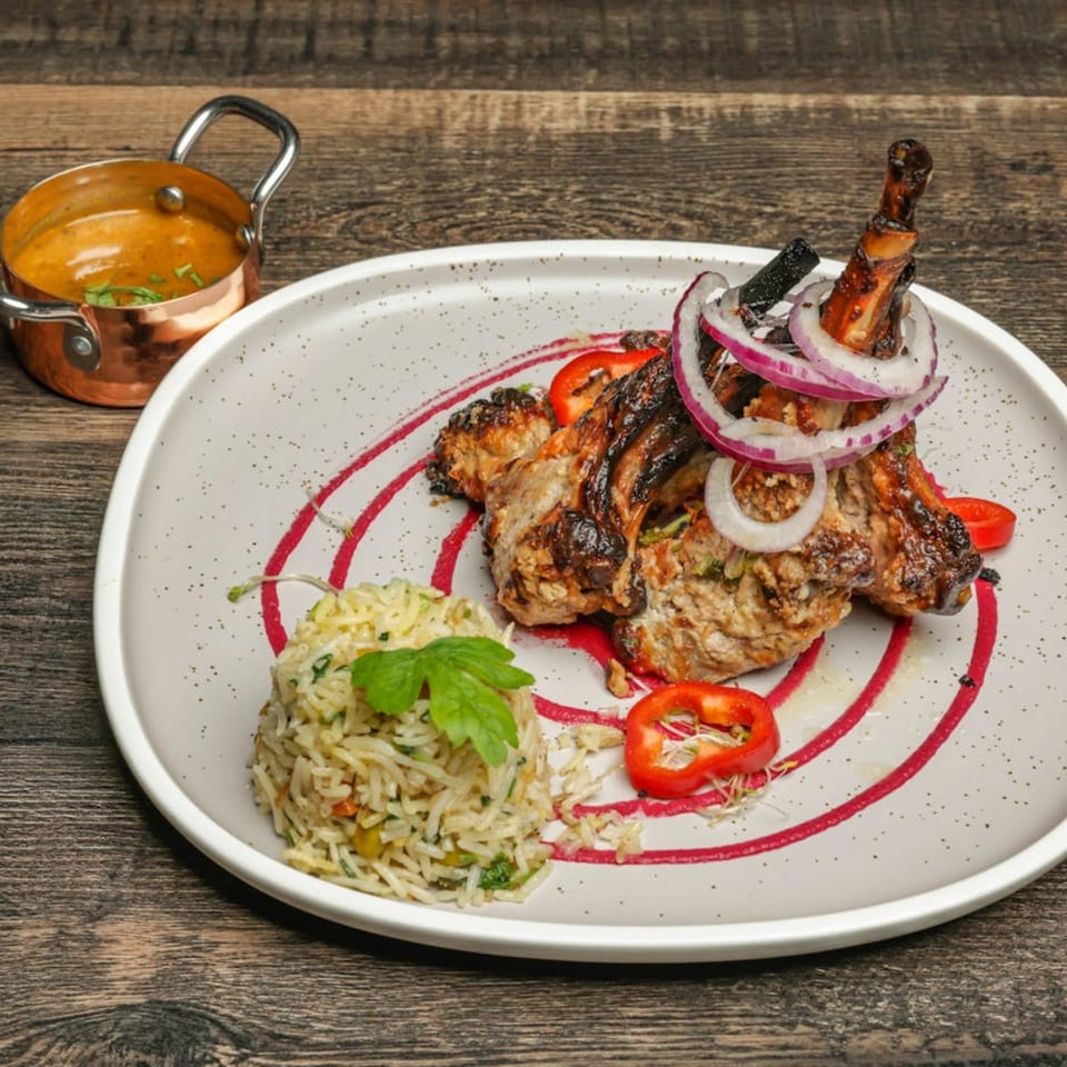 Grilled lamb chops with red onion, rice, and a side of curry on a white plate.