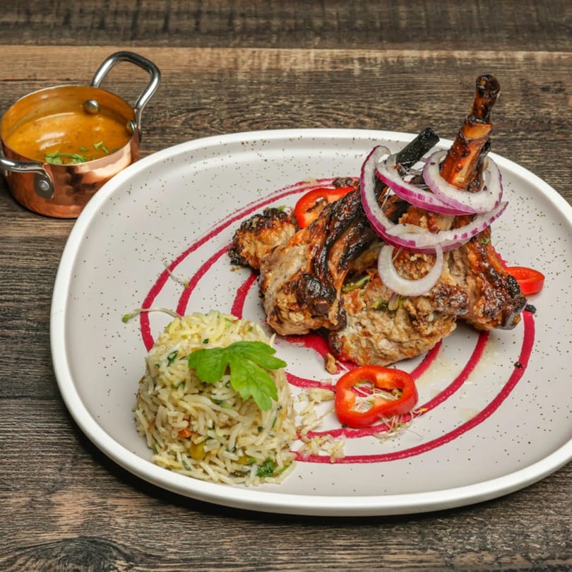 Grilled lamb chops with red onion, rice, and a side of curry on a white plate.