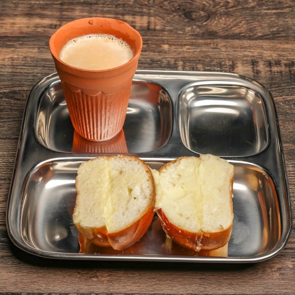 Mumbai Maskaa's chai in a clay cup and buttered pav bread on a metal tray.