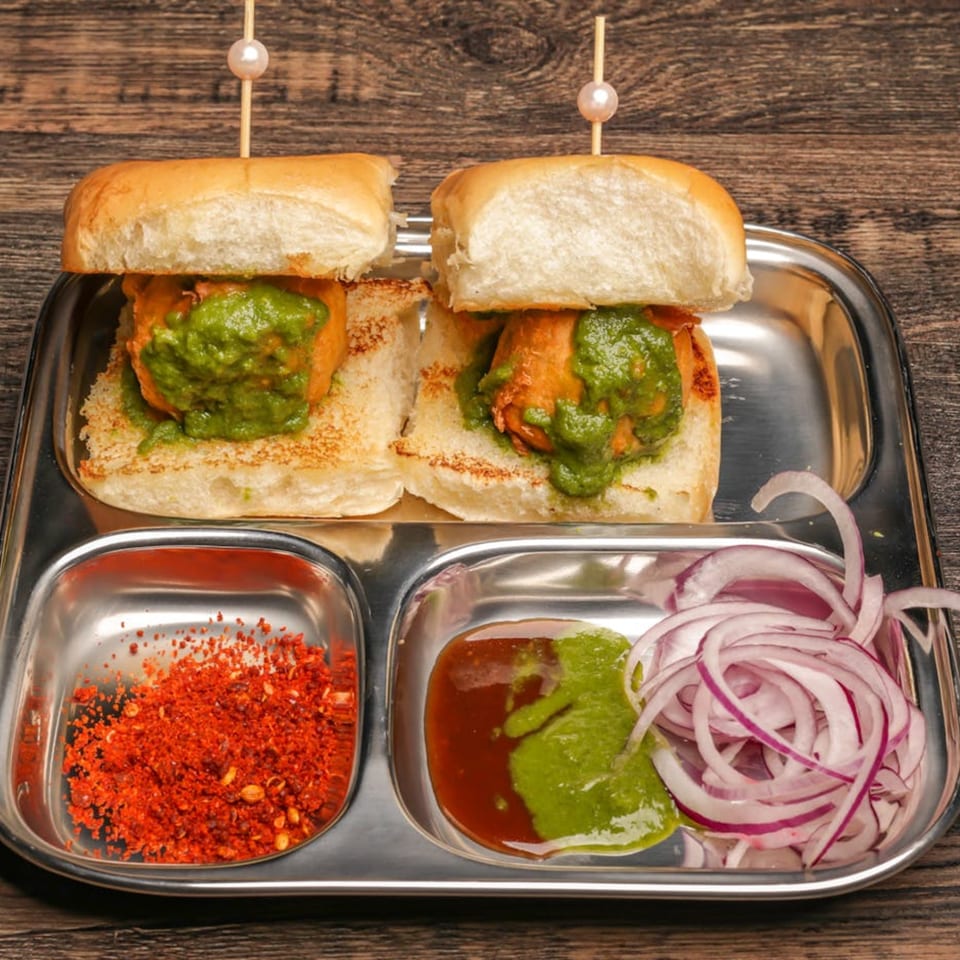 Two Mumbai Maskaa vada pavs with green chutney, red chili powder, and sliced red onions.