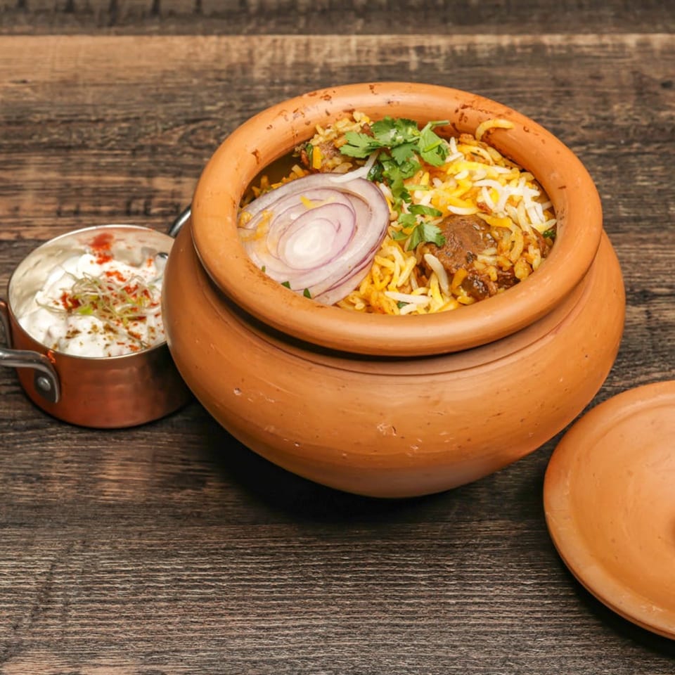 Indian biryani in a clay pot with sliced onions, cilantro, and a side of raita.