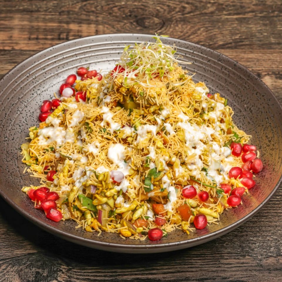 A vibrant Mumbai Maskaa Bhel Puri, topped with sprouts, pomegranate seeds, and white sauce, on a dark plate.