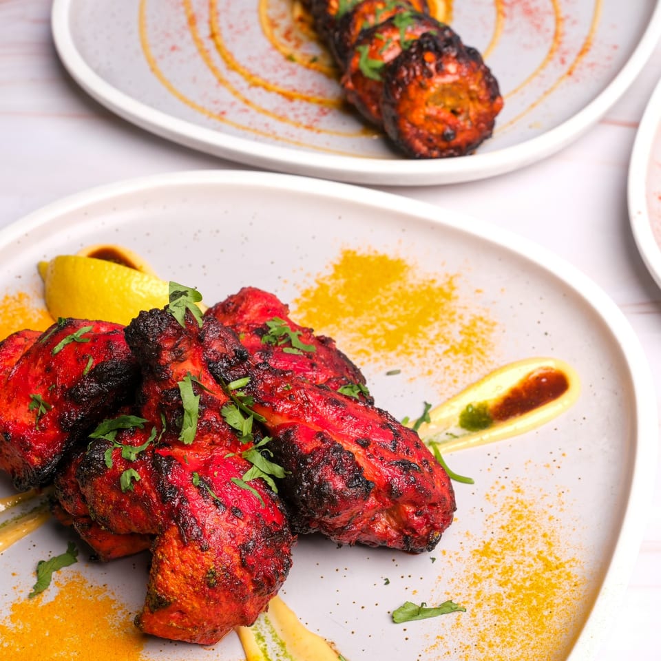 Vibrant red tandoori chicken drumsticks with cilantro, lemon, and sauces on a light gray plate.