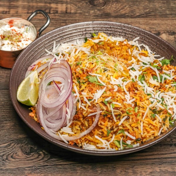 Indian biryani with rice, onions, lime, and a side of raita on a wooden table.