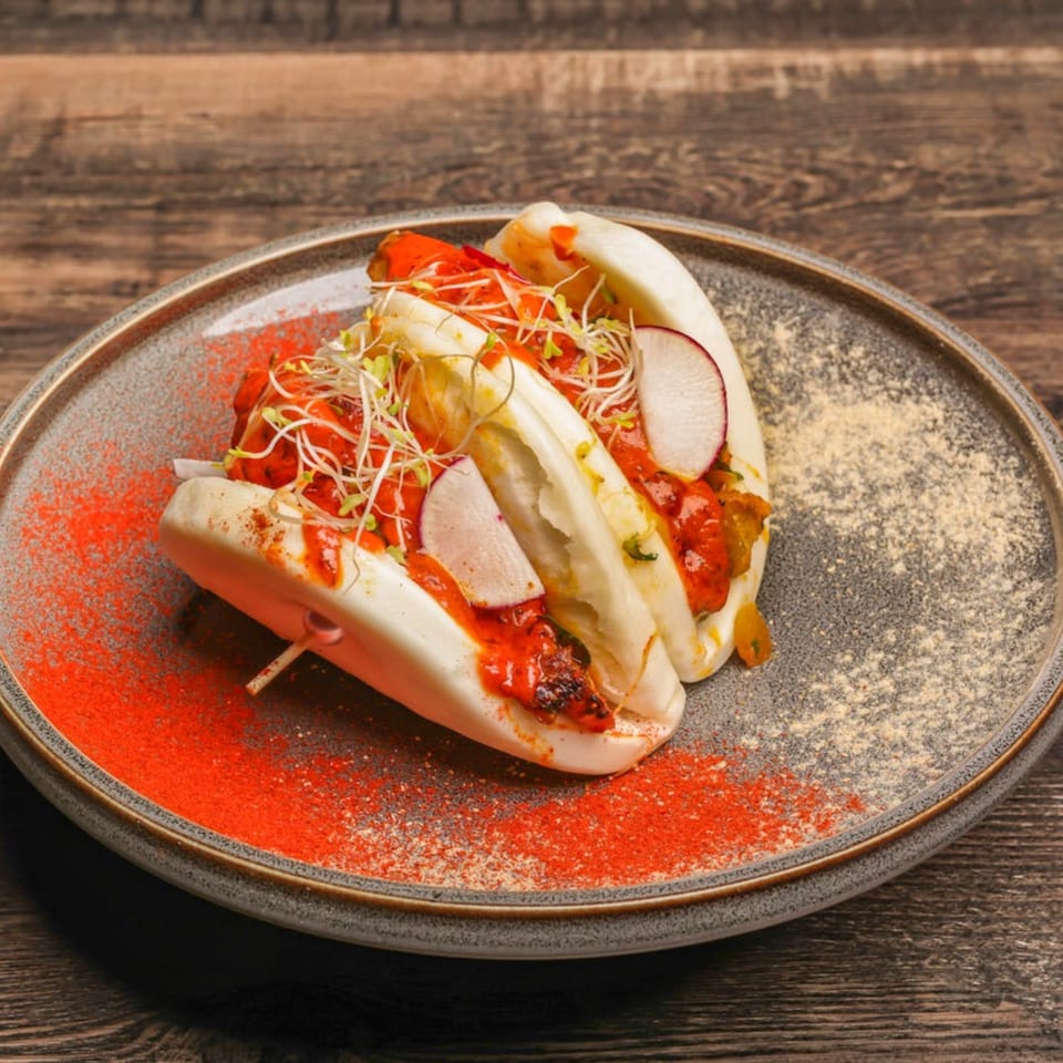 Two Mumbai Maskaa Indian bao buns with red sauce, radishes, and sprouts on a plate.